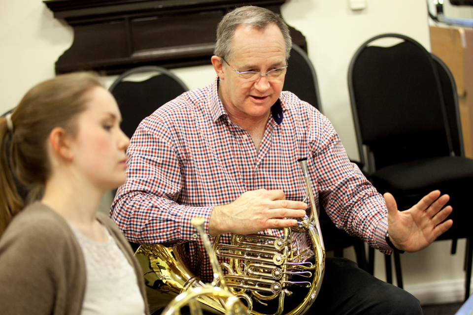 A male professor, with light hair and glasses, holding a french horn, instructing a female student, holding a french horn, in a 1-2-1 lesson.